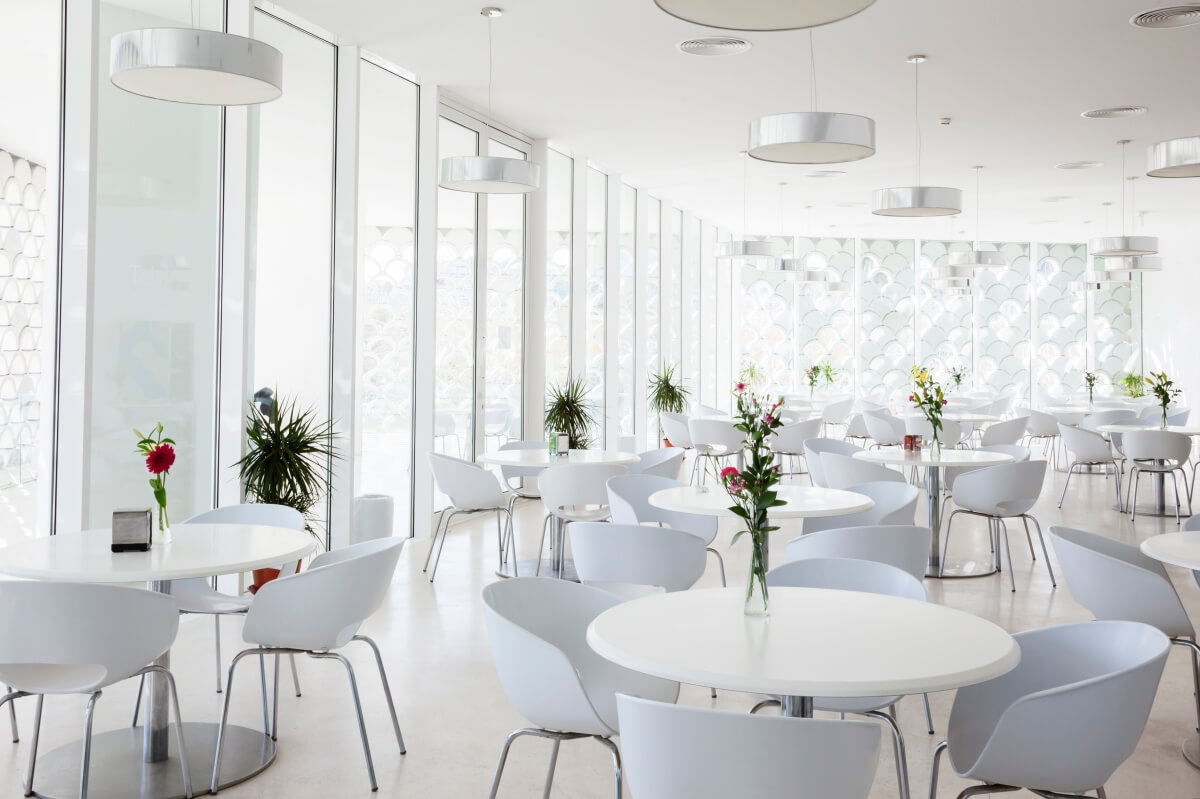 Nowoczesna jadalnia z białymi krzesłami i stołami, gdzie podstawy do stołów okrągłe, a na nich stoją wazoniki z kwiatami.