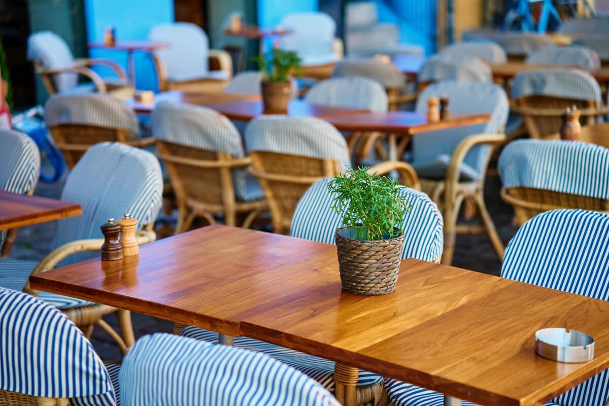 Na zewnątrz stoją drewniane stoły i krzesła ogrodowe z poduszkami w paski.